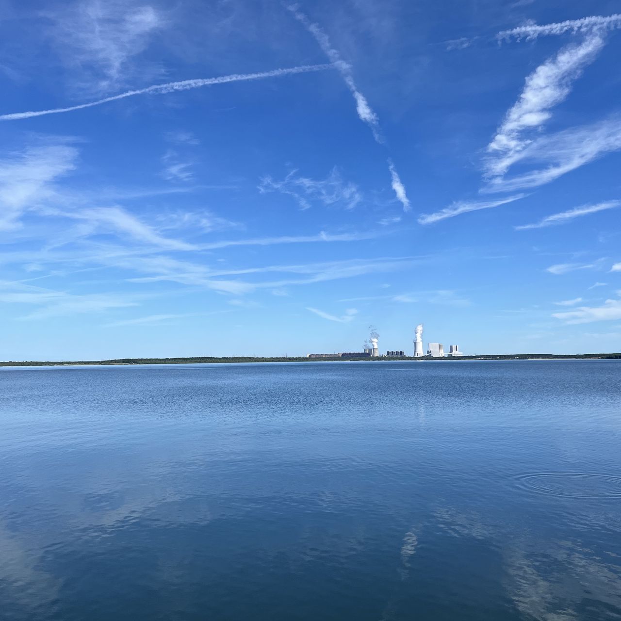 A color foto of a lake with a coal power plant in the distance and a blue sky with some clouds and traces of planes.